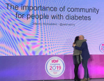 Me giving Manny Hernandez a hug. We are standing on a stage with a purple background which is a giant screen. The words 'The importance of community for people with diabetes' (and Manny's name and Twitter handle @AskManny) is written in black above us. The logo for the 2019 IDF Congress is also on the screen. 